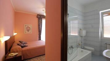 Un dormitorio con cama, mesita de noche y ventana con cortinas. Junto a él, un baño con bañera, inodoro y lavabo.