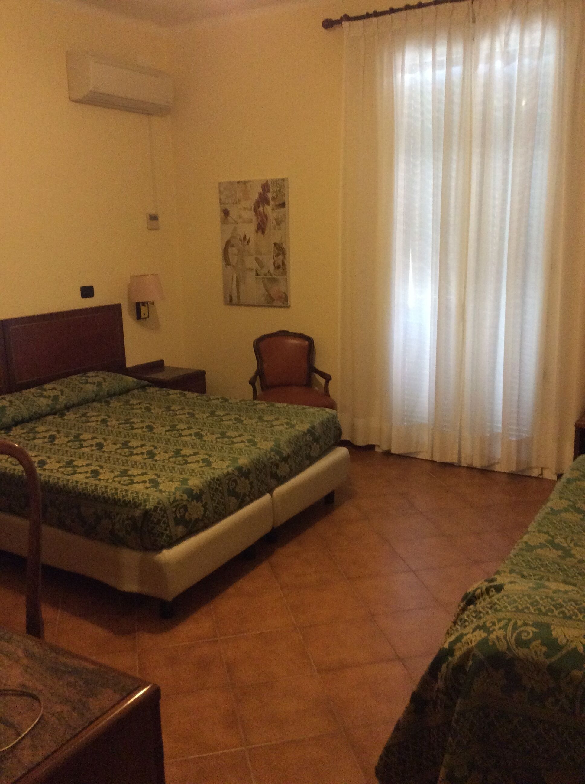 Una habitación de hotel con una cama, una silla, una mesa, una lámpara, un cuadro y una ventana con cortinas.