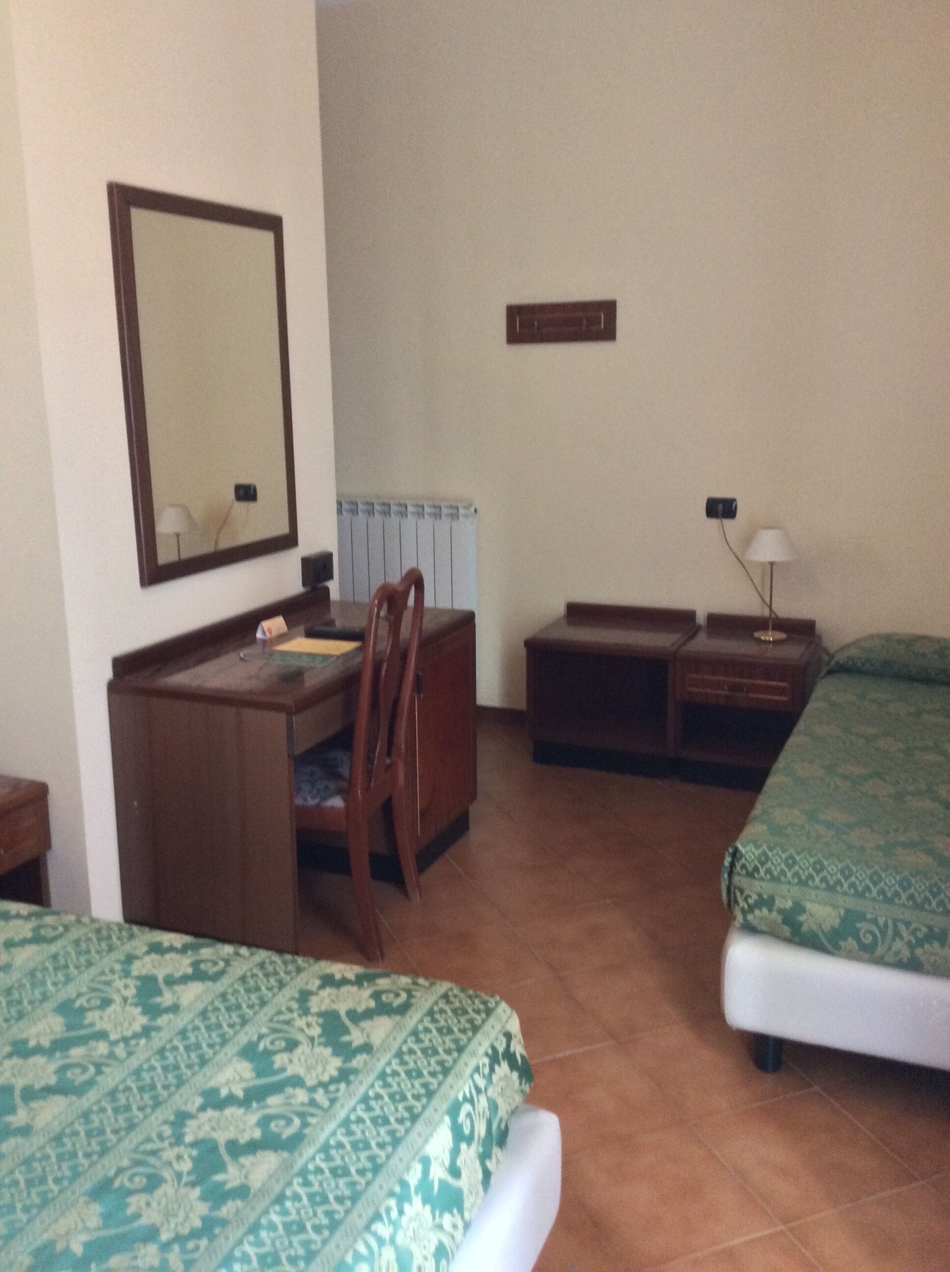 A hotel room with a bed, a desk, a chair, a nightstand, a lamp, and a mirror.