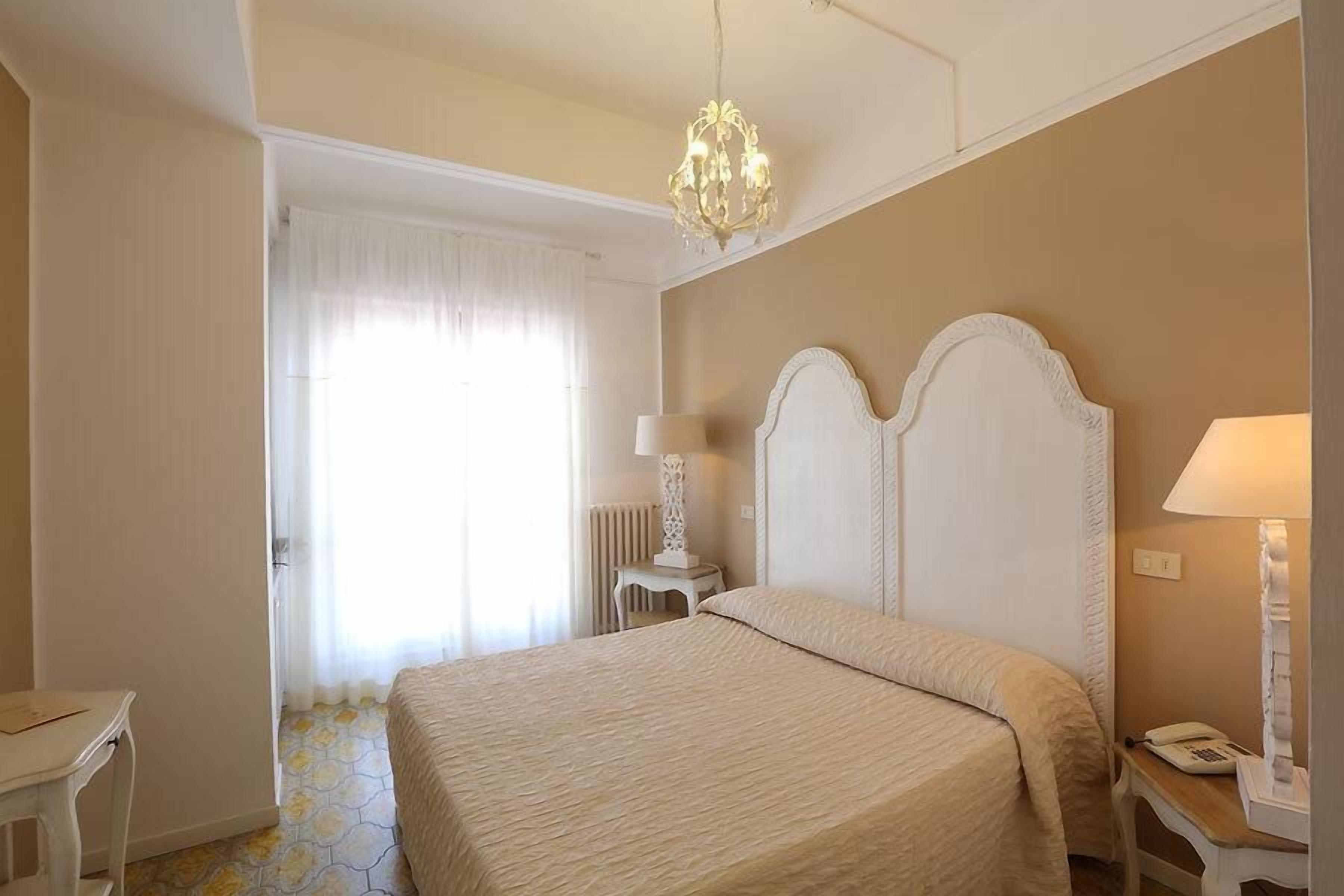 Una habitación de hotel con una cama grande, una araña, una mesita de noche con lámpara y un ventanal con cortinas.