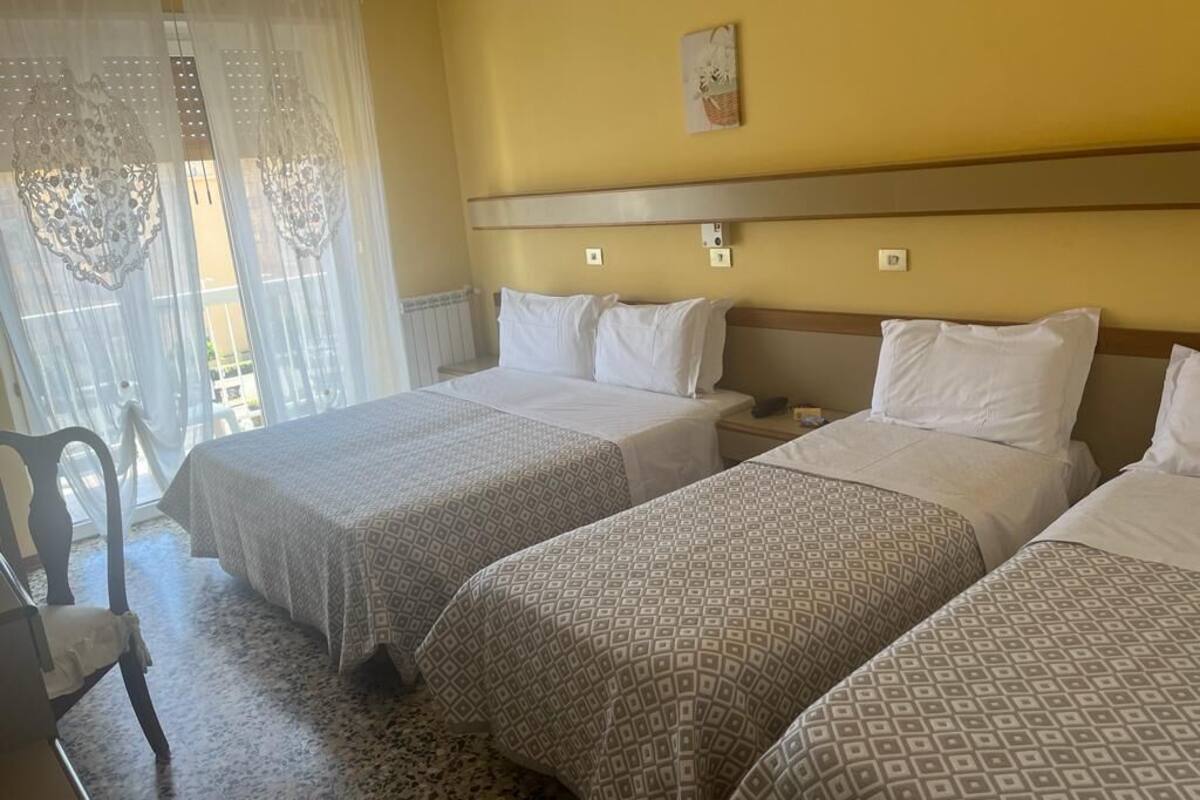 Una habitación de hotel con dos camas individuales, una silla, una mesita de noche y una ventana con cortinas translúcidas.