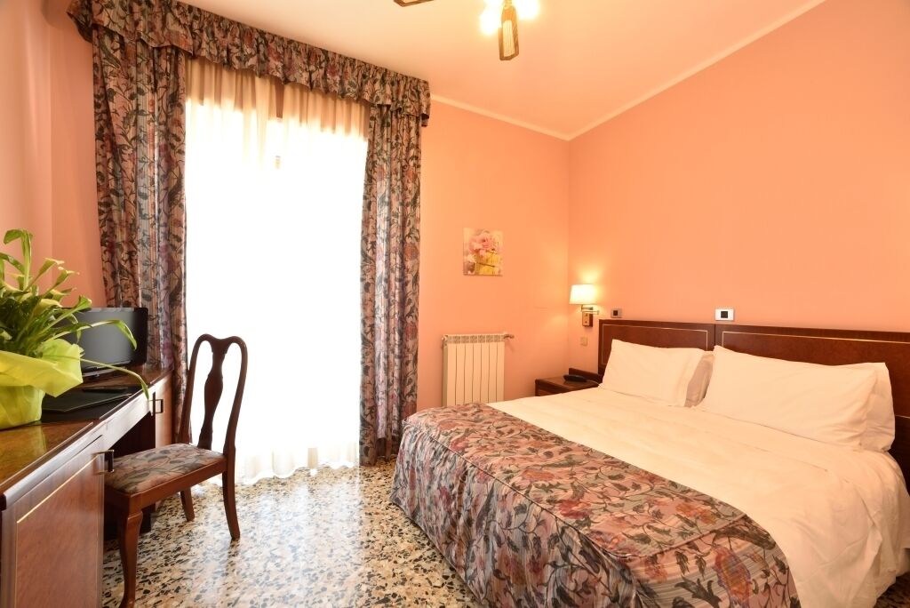 A hotel room with a bed, a desk with a chair, a radiator, and a wall-mounted light.