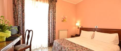 A hotel room with a bed, a desk with a chair, a radiator, and a wall-mounted light.