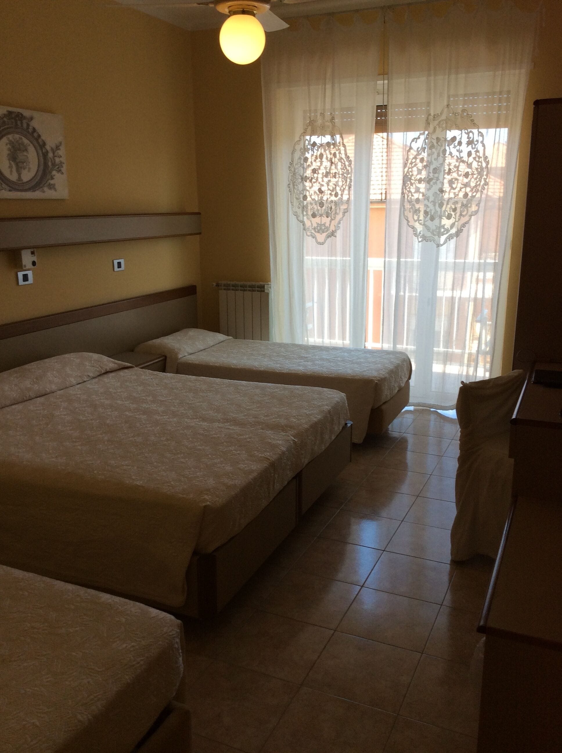 Habitación de hotel con dos camas, ventilador de techo, radiador y ventana con cortinas ligeras.