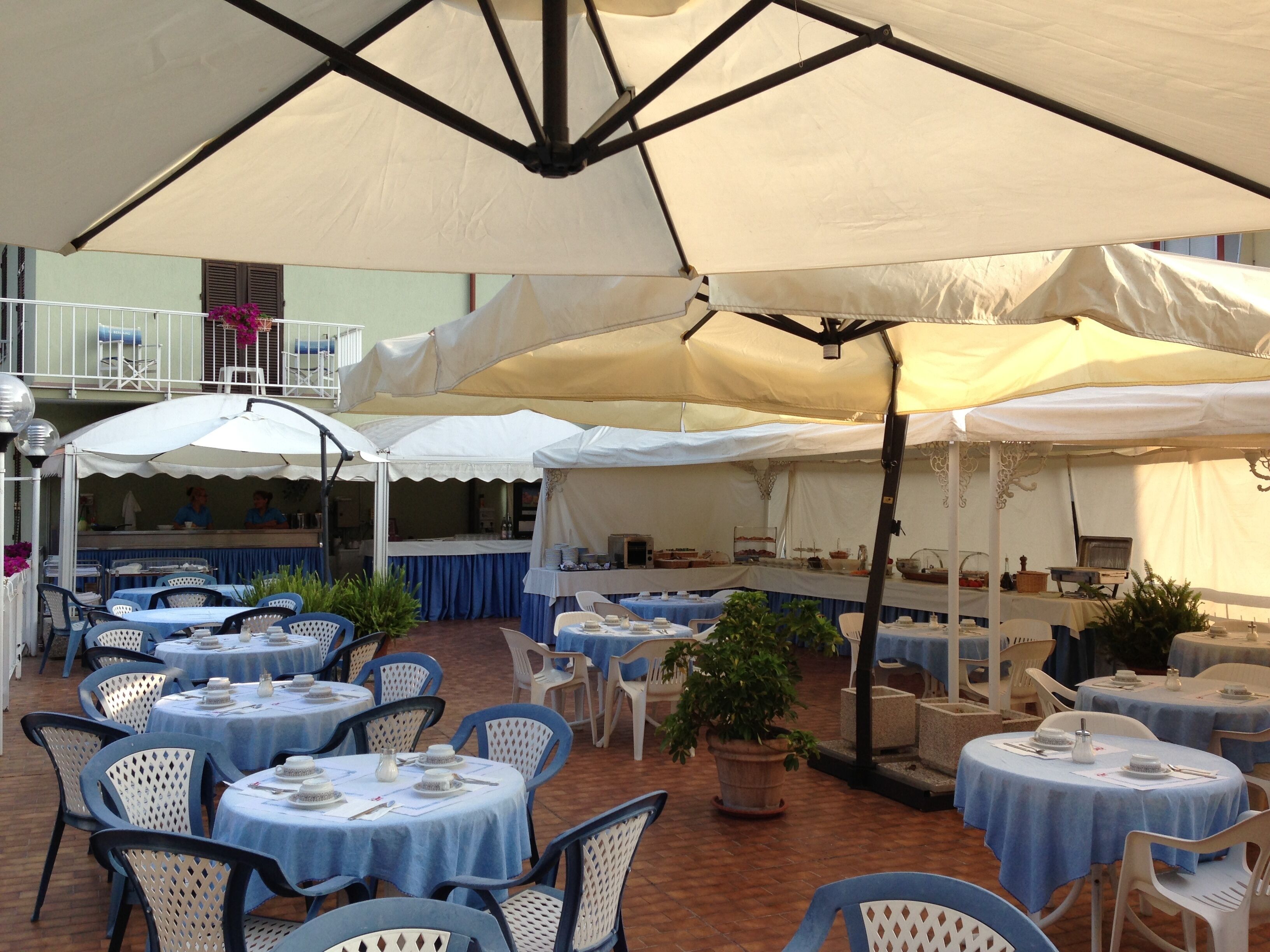 An outdoor dining area with round tables covered by blue tablecloths and white umbrellas, surrounded by white chairs.