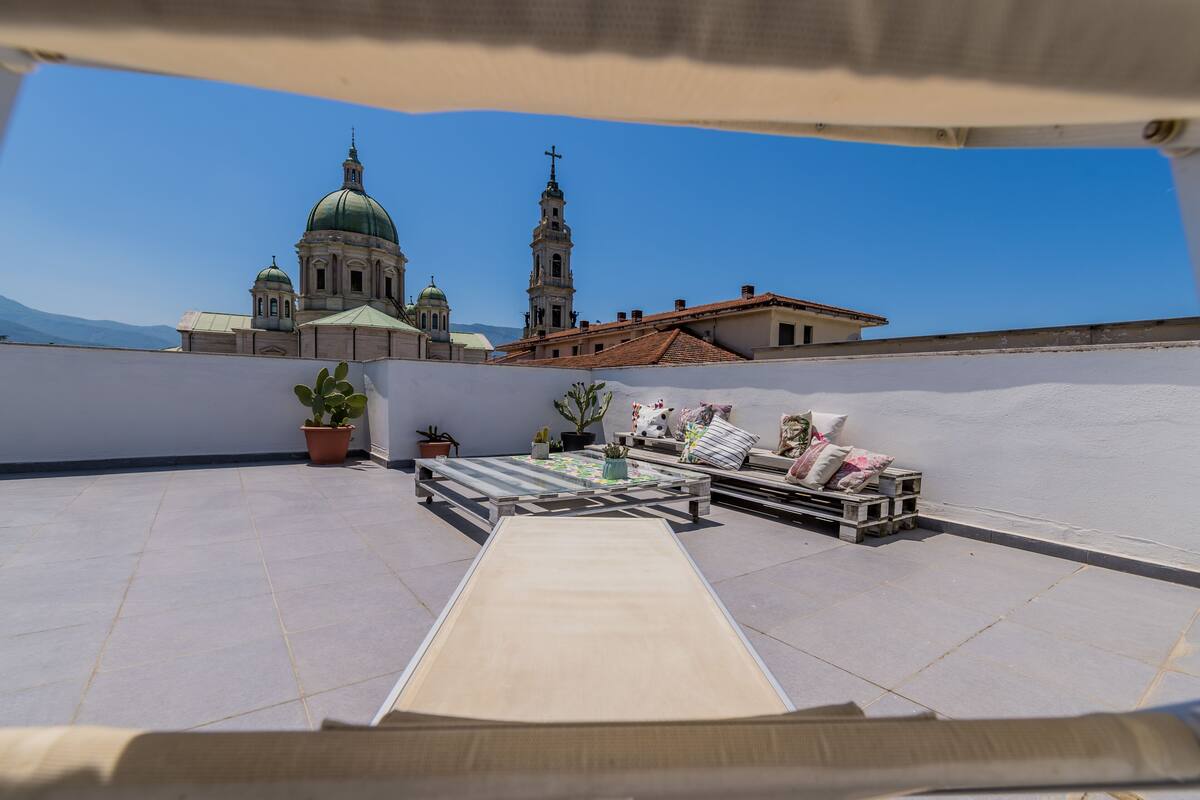 Una terraza en la azotea con una mesa de comedor y sillas, plantas en macetas y un sofá. Al fondo, se ve una iglesia grande con dos torres y una cúpula.