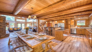 A spacious log cabin interior with a long dining table, a cozy seating area, a kitchen, and a staircase leading to an upper level.