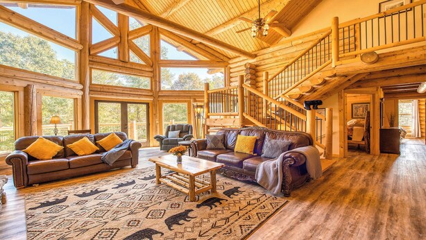A spacious log cabin interior with a large living area, a wooden staircase, and large windows.
