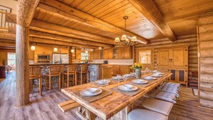 A log cabin interior with a long dining table set for six, wooden bar stools, a stone accent wall, and a kitchen area with modern appliances.