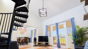 Uma sala de estar espaçosa com escadas, uma TV de tela plana em um suporte, área de jantar com cadeiras e mesa, e janelas grandes com cortinas.
