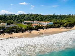 A beachfront property with a large building, a sandy beach, and a forested hill in the background.