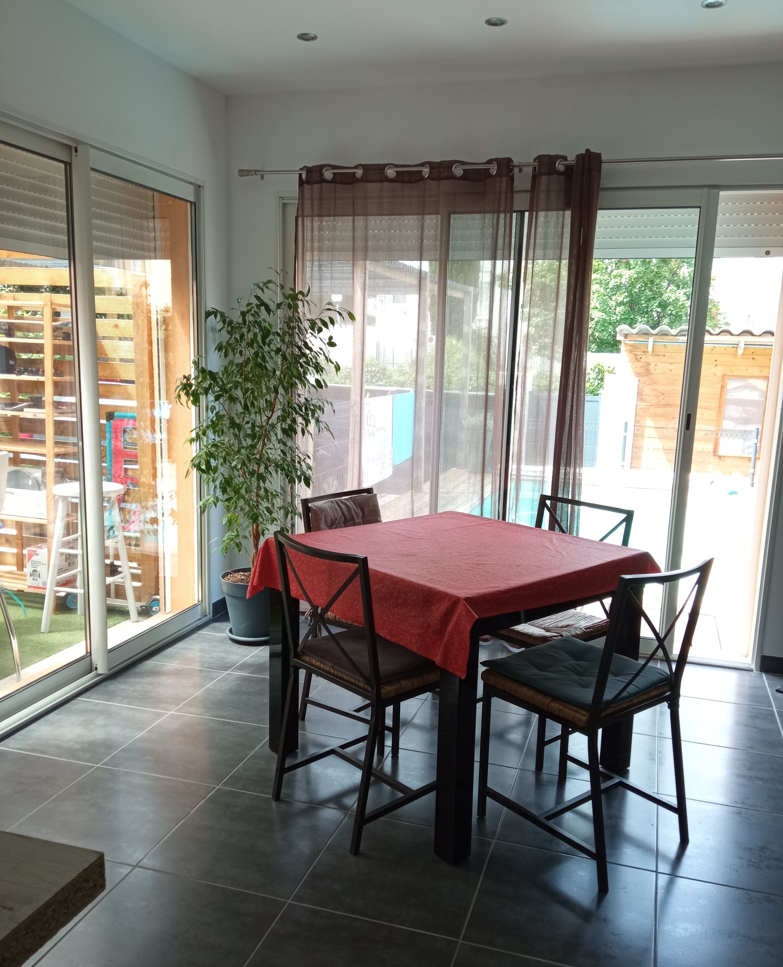 A dining area with a table covered in a red tablecloth, surrounded by four chairs. There is a potted plant to the left of the table.