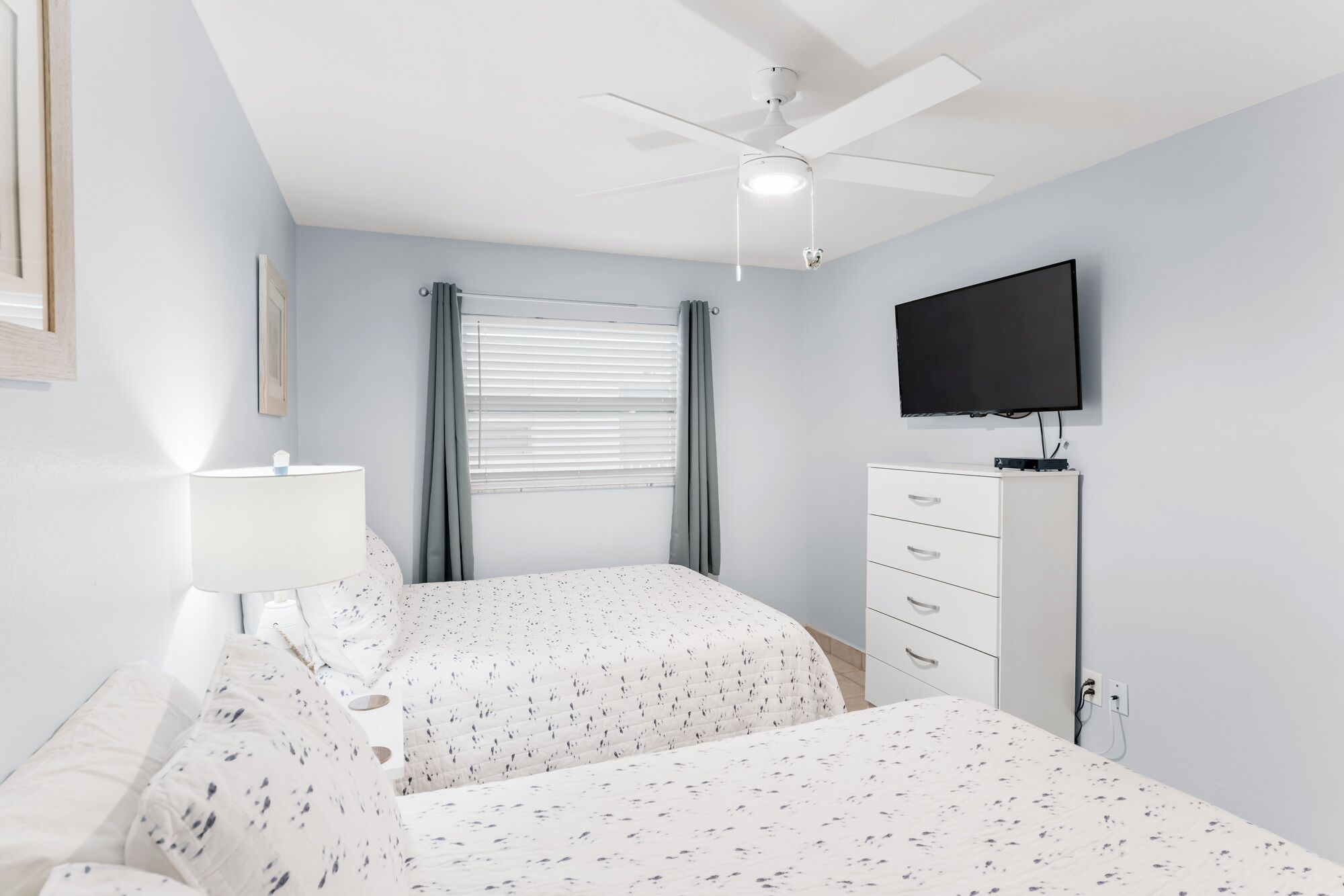 A bedroom with two beds, a ceiling fan, a TV, a dresser, and a window with curtains.