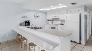 Una cocina moderna con armarios blancos, una isla central con taburetes, microondas, cafetera y un reloj en la pared.