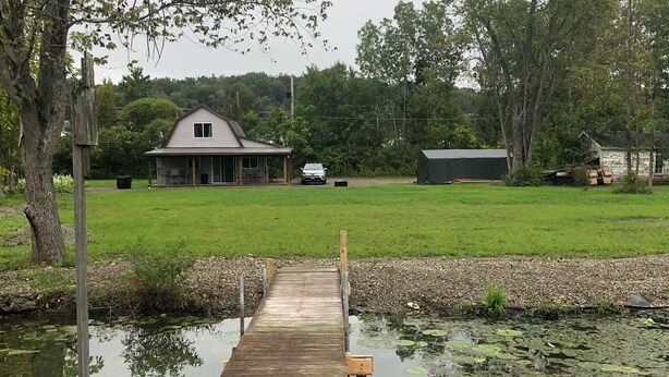 A wooden dock extending into a calm body of water with lily pads, surrounded by trees and a grassy area with a house in the background.