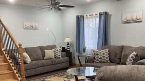 A living room with a wooden staircase, two grey sofas, a glass coffee table, and a ceiling fan.