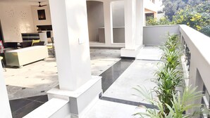 Un espacio exterior moderno con una zona de estar, un mostrador de recepción blanco y un balcón con plantas en macetas.