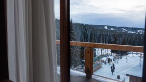 A view from a window with a wooden balcony overlooking a snowy landscape with trees and a ski slope.