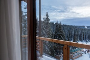 A view from a balcony with a wooden railing, overlooking a snow-covered forest and a building with outdoor seating. - Hotel Rtanj Kopaonik (Raška)