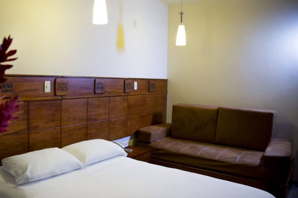 Quarto de hotel com cabeceira de madeira, cama com lençóis brancos, sofá marrom e duas luminárias pendentes.