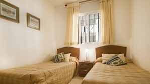Ein Zimmer mit zwei Betten, einem Nachttisch, einem Fenster mit Vorhängen und gerahmten Bildern an der Wand.