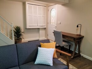 A living room with a gray sofa, a wooden desk, and a chair. There is a potted plant and a white door with a window above it. - Spacious 800 sqf 1BR, 2 minutes to subway + 6 minutes to Midtown Manhattan (Queens)