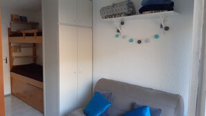 A room with a bunk bed, a sofa with blue cushions, a wall-mounted shelf with books, and a decorative string of round objects. - Studio Chaleureux en Montagne (Puyvalador)