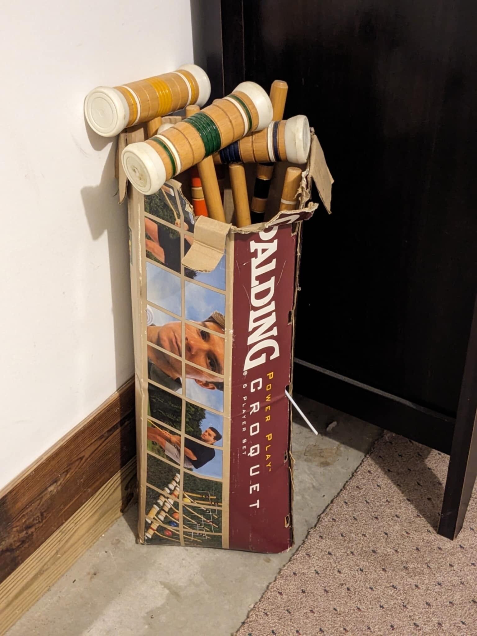 Una caja de cartón etiquetada como 'Palding Croquet Set' con una colección de mazas y pelotas de croquet.