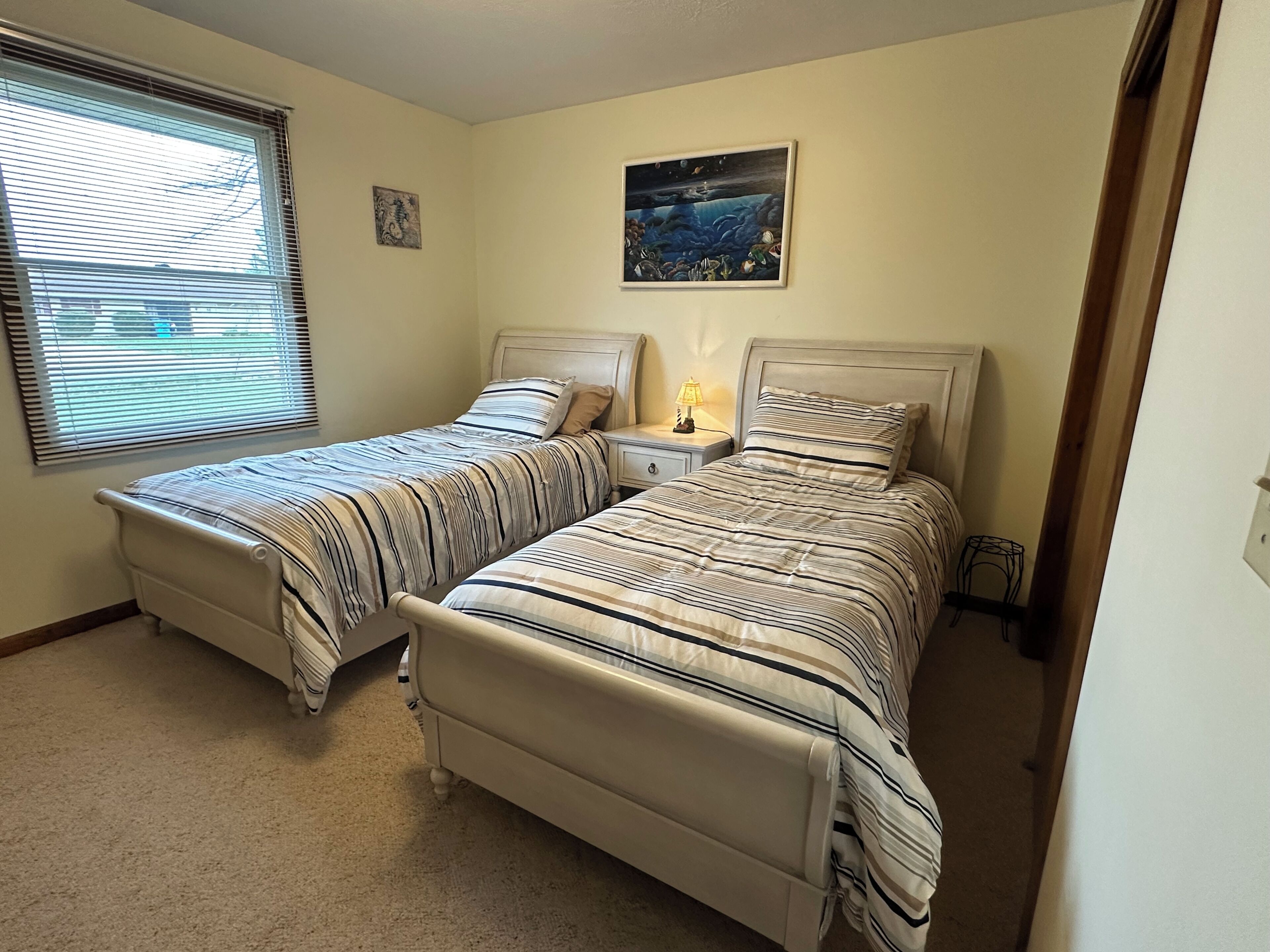 A bedroom with two beds, a nightstand with a lamp, a painting on the wall, and a window with blinds.