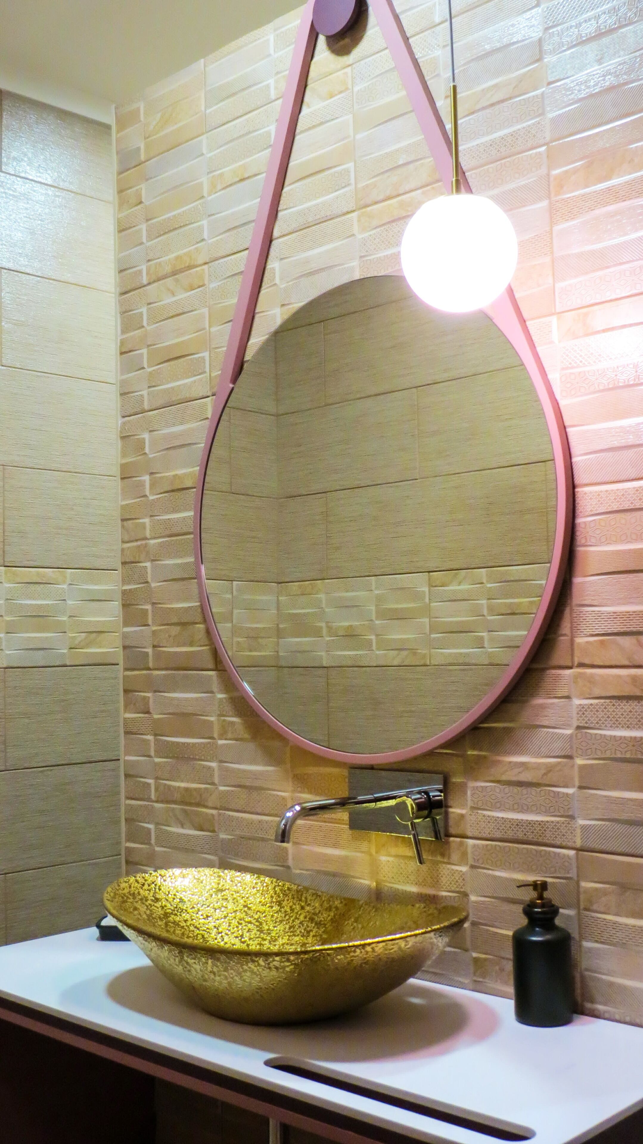 A bathroom with a round mirror, a gold bowl sink, and a pink light fixture.