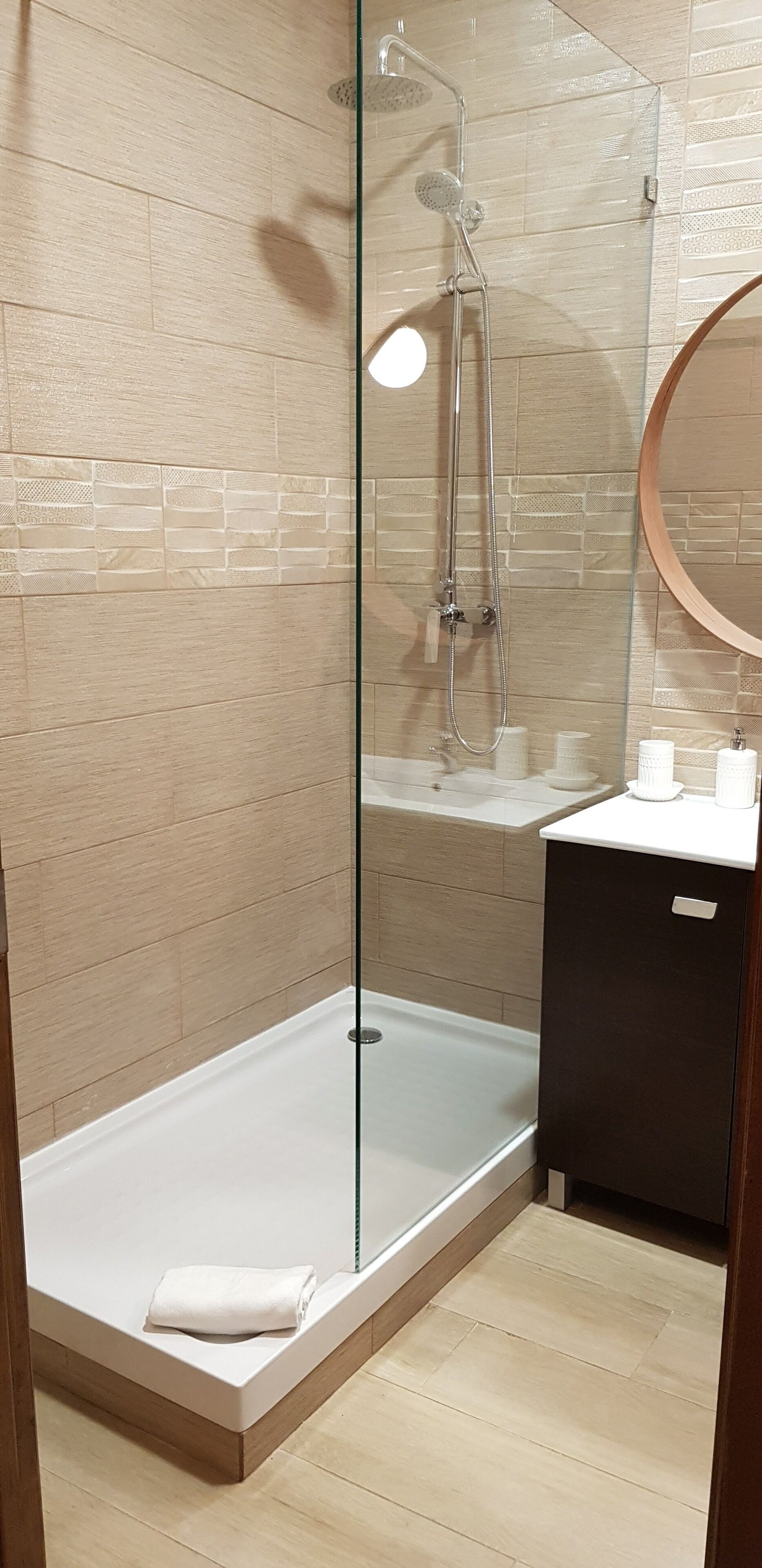 Un baño moderno con ducha acristalada, suelo de madera y un mueble oscuro pequeño.