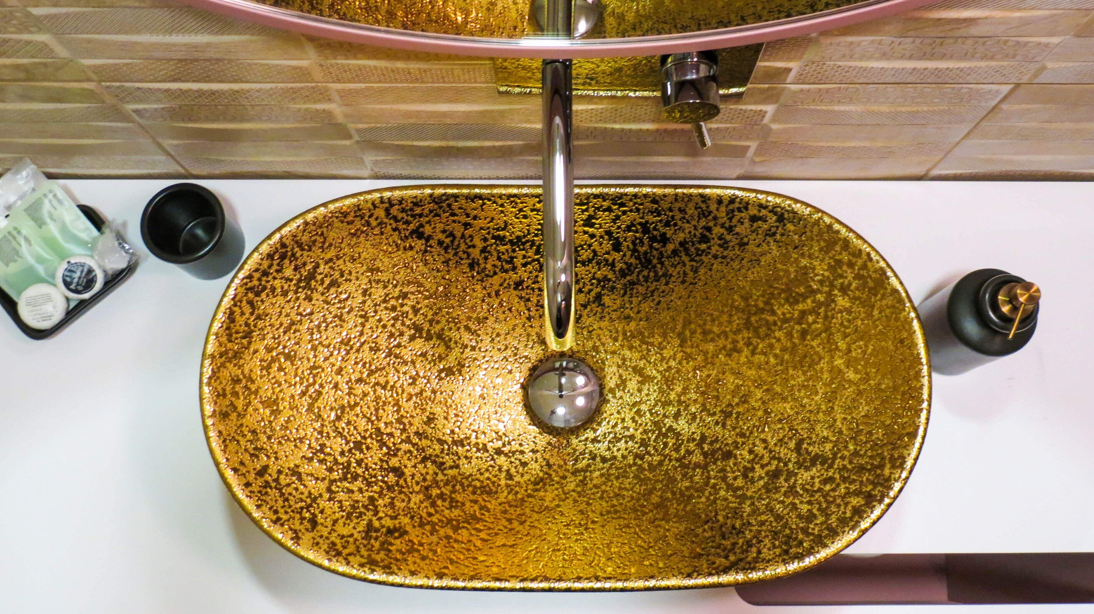 Un lavamanos dorado con un grifo moderno, una taza pequeña y una botella de jabón para manos en la encimera.