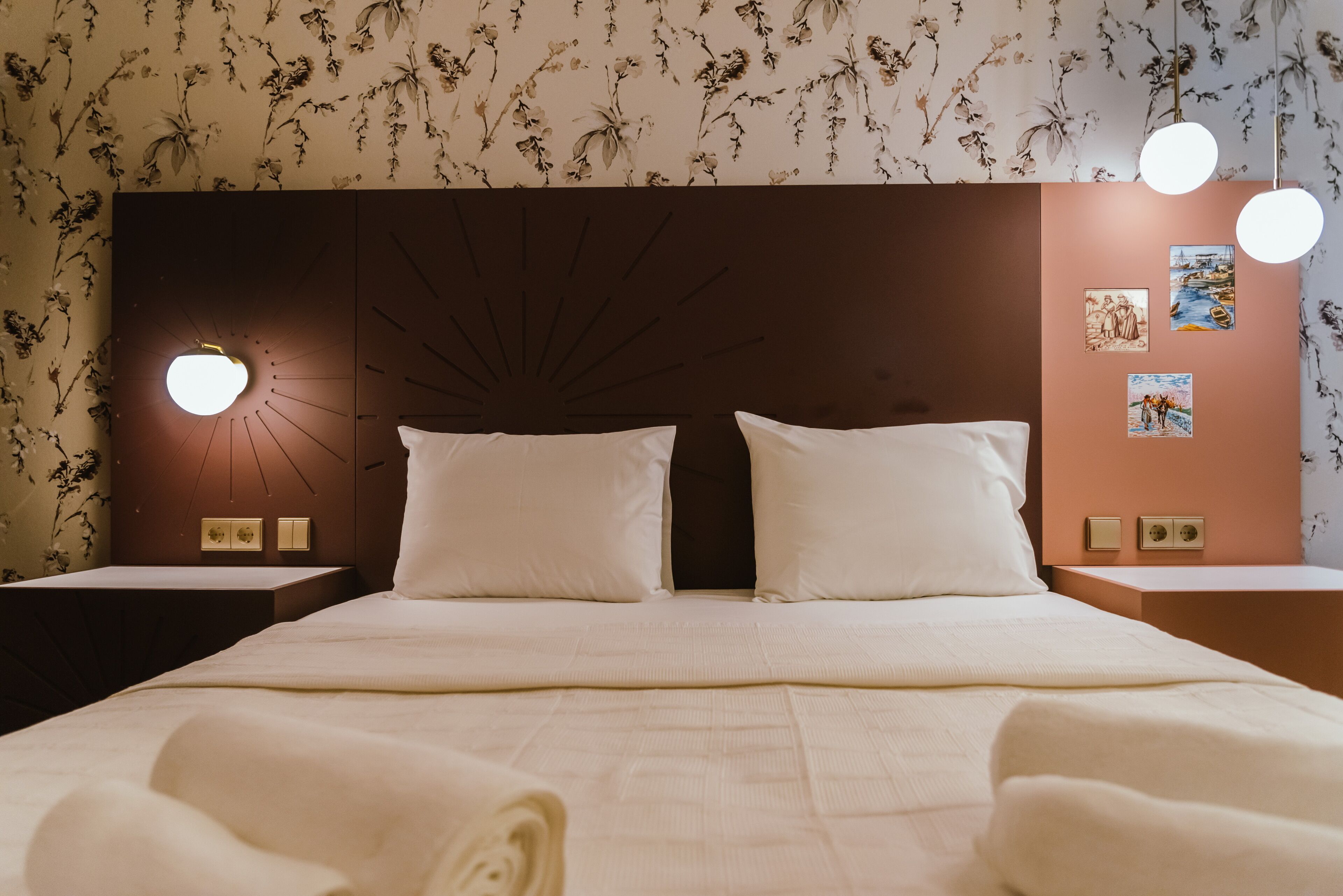 A neatly made bed with white linens, a wall-mounted round light, and floral wallpaper.