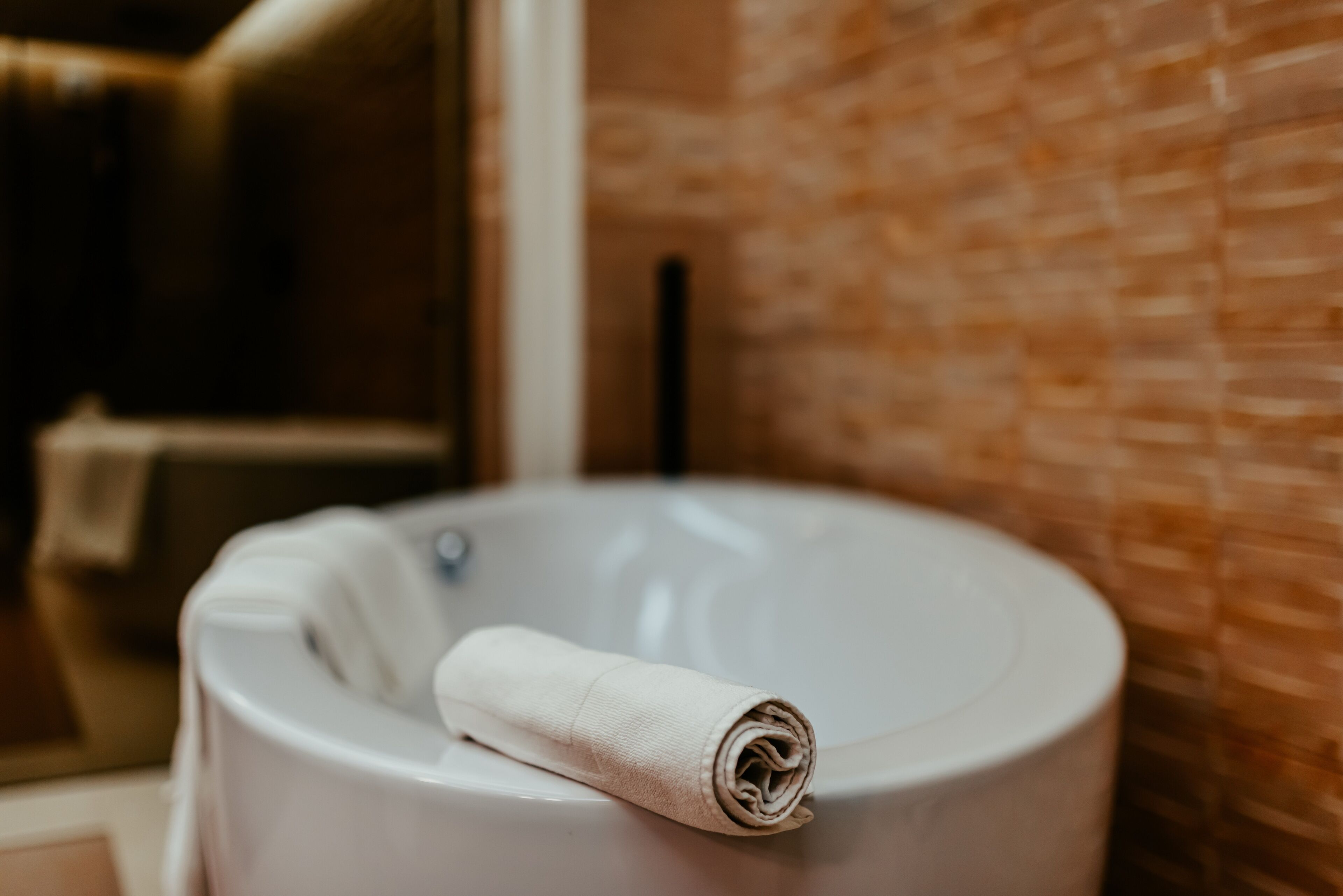 Eine weiße Badewanne mit einem aufgerollten Handtuch darauf, einem sichtbaren Wasserhahn und einer strukturierten Wand im Hintergrund.