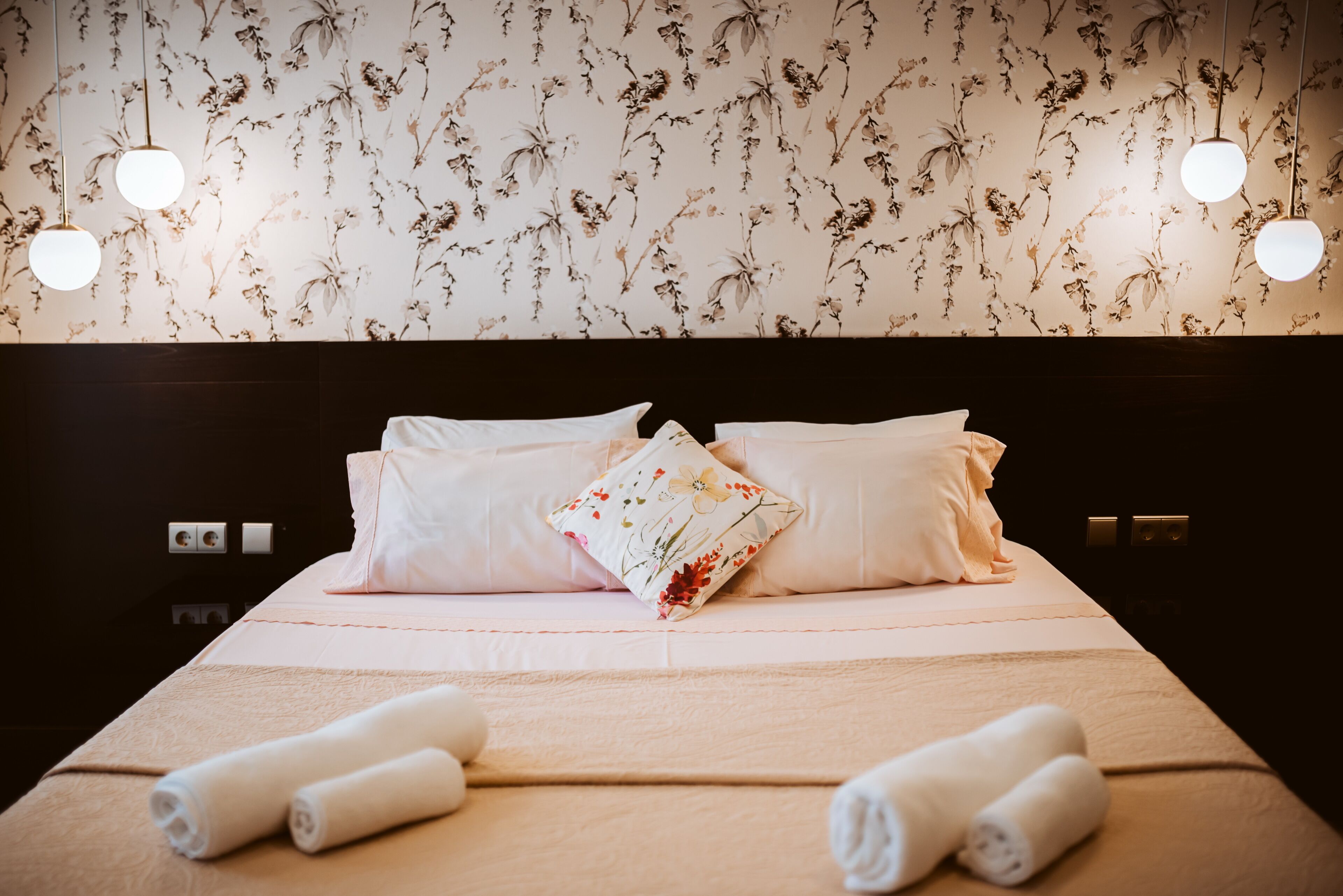 A neatly made bed with white linens and floral-patterned pillows, two wall-mounted light fixtures, and a floral wallpaper.