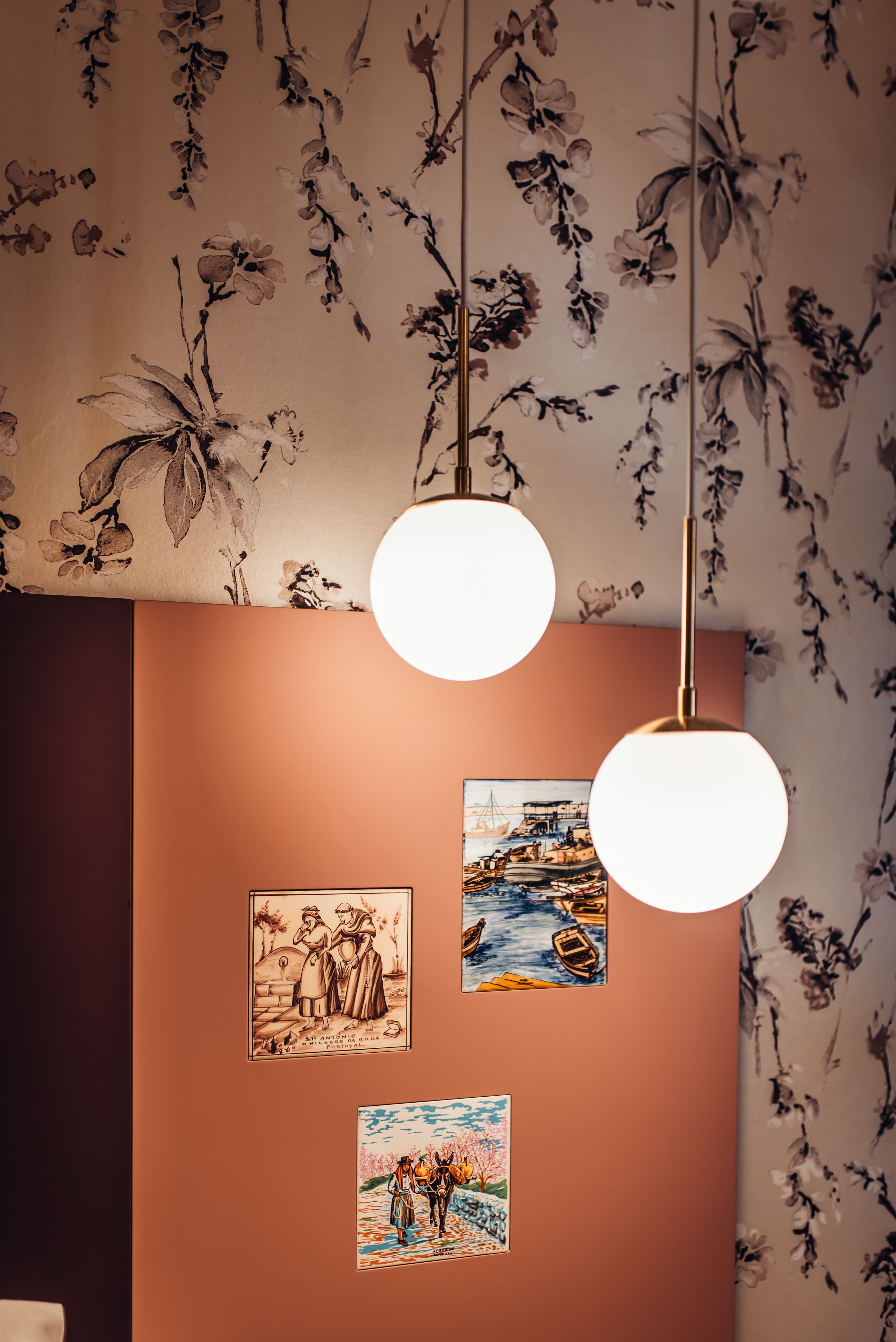 Une pièce dont les murs sont recouverts d’un papier peint à motifs floraux, avec deux suspensions rondes au plafond et trois œuvres d’art encadrées accrochées au mur.