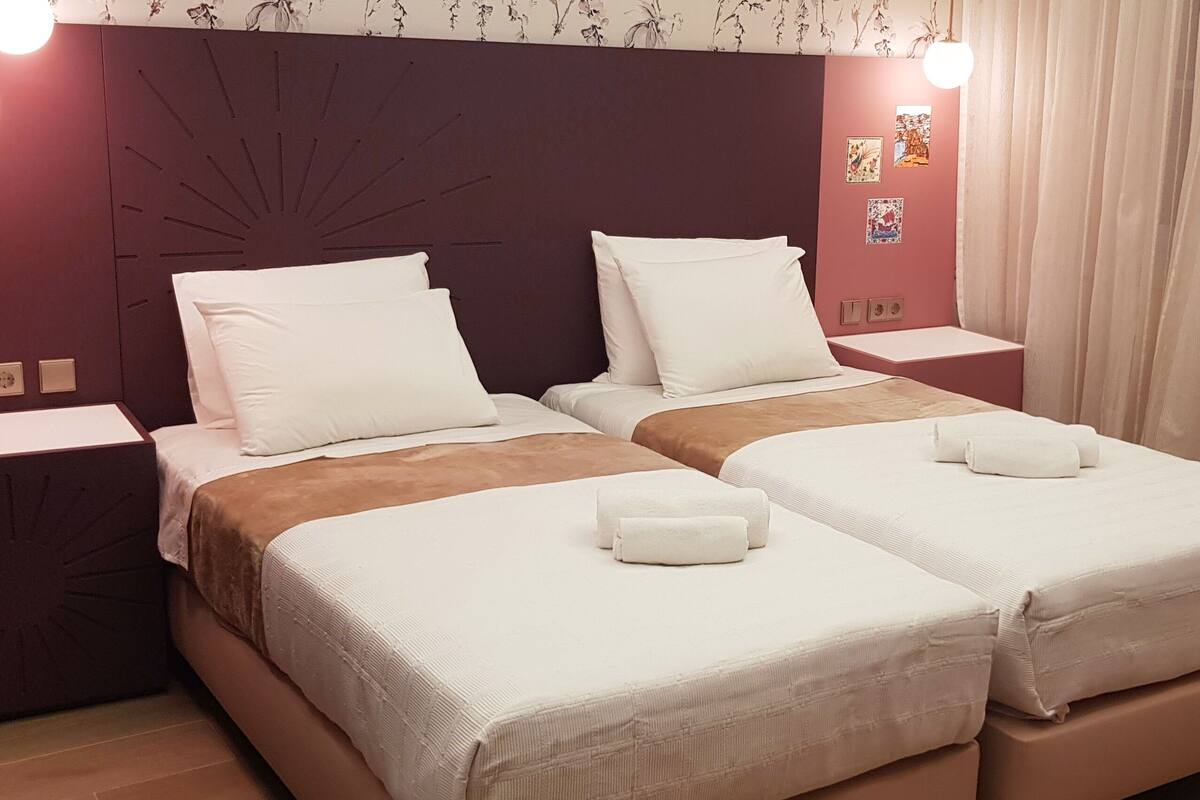 A hotel room with two beds, each with white linens and a brown blanket. There are two bedside tables, a headboard with a sunburst design, and a wall with framed pictures.