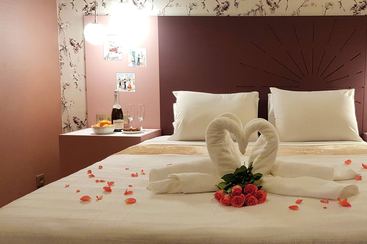 A hotel room with a bed featuring a heart-shaped towel arrangement, a bouquet of roses, and rose petals scattered on the bed.