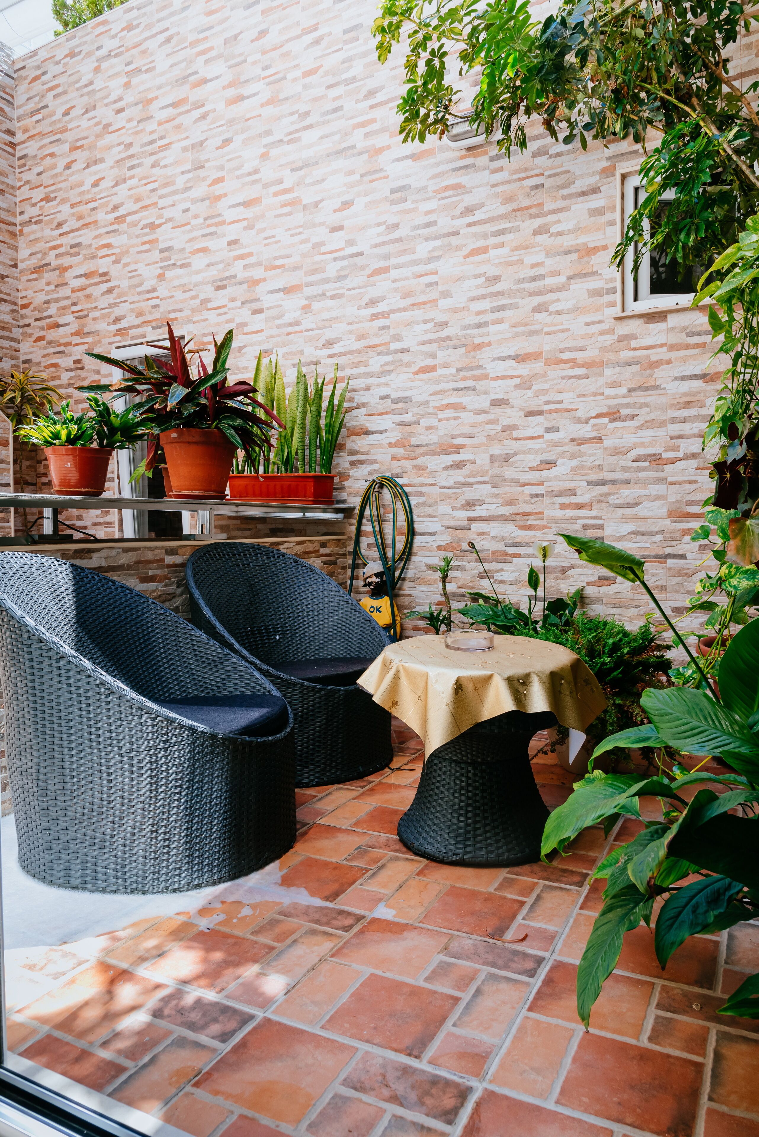 A patio area with wicker furniture, a small table, and potted plants against a brick wall.