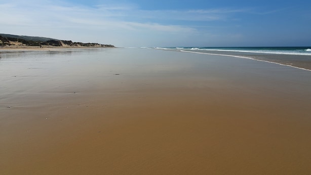 Ein weiter Sandstrand mit sanften Wellen, blauem Himmel und Hügeln im Hintergrund.