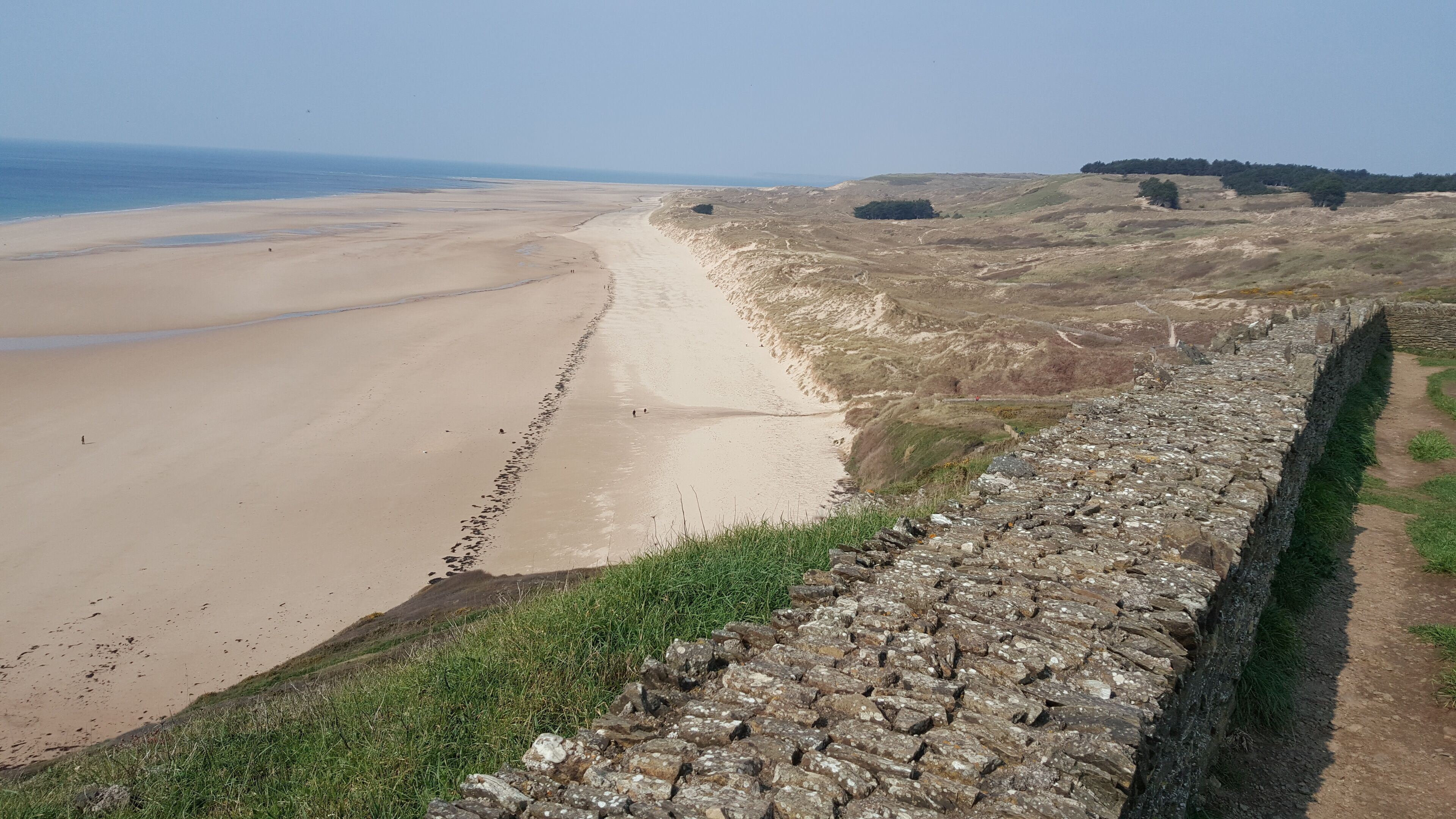 A coastal landscape with a sandy beach, a stone wall, and a grassy hill.