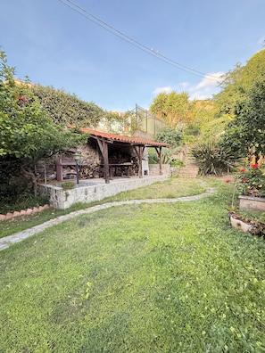 A rustic outdoor area with a stone bench, a wooden table, and a small wooden structure with a tiled roof.