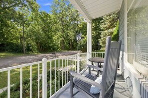 Eine Veranda mit Bank und Tisch, umgeben von einem weißen Geländer, mit Blick auf eine Straße und dichtes Grün.