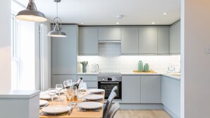 A modern kitchen with grey cabinets, a built-in oven, and a dining area set for a meal. - The Escape (Brighton)