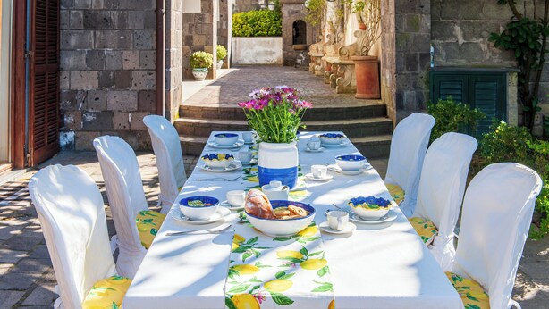 A table set for a meal with chairs covered in white cloth, a vase with flowers, and a tablecloth with a lemon pattern. - Beautiful Villa with Private Pool and Garden (Massa Lubrense)