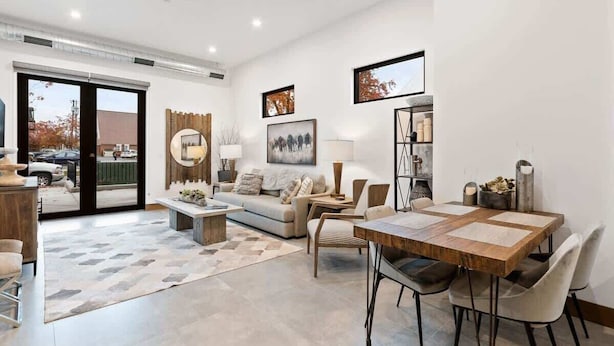 A modern living room with a sofa, coffee table, dining table, and chairs. There is a large window with a view of the outside.