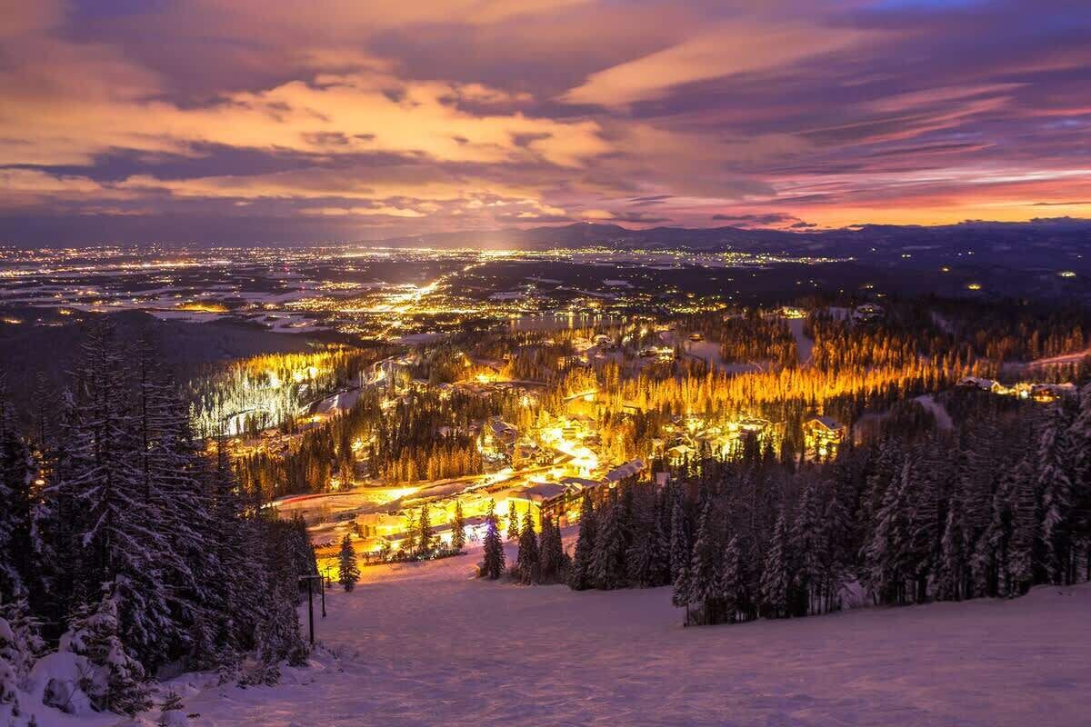 A snow-covered landscape with a town illuminated by lights, surrounded by trees and a clear sky.