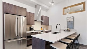 A modern kitchen with stainless steel appliances, a central island with bar stools, and a hanging light fixture.