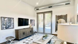 A modern living room with a flat-screen TV, a wooden cabinet, a dining table, and a large window.