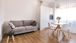 Un salon moderne avec un canapé gris, des chaises blanches, une table à manger en bois et un coin cuisine doté de meubles blancs et d’un four.
